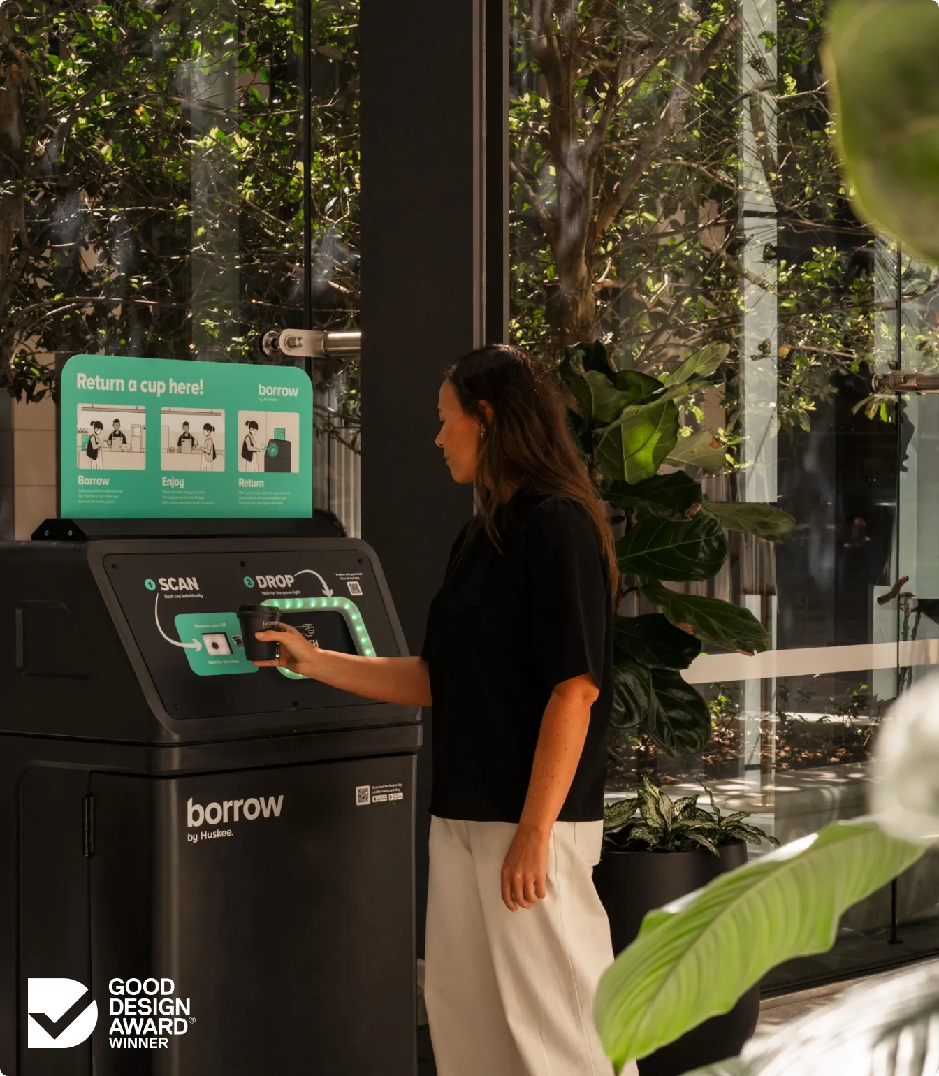 A woman returning a cup at a Borrow bin between a glass wall and potted plants