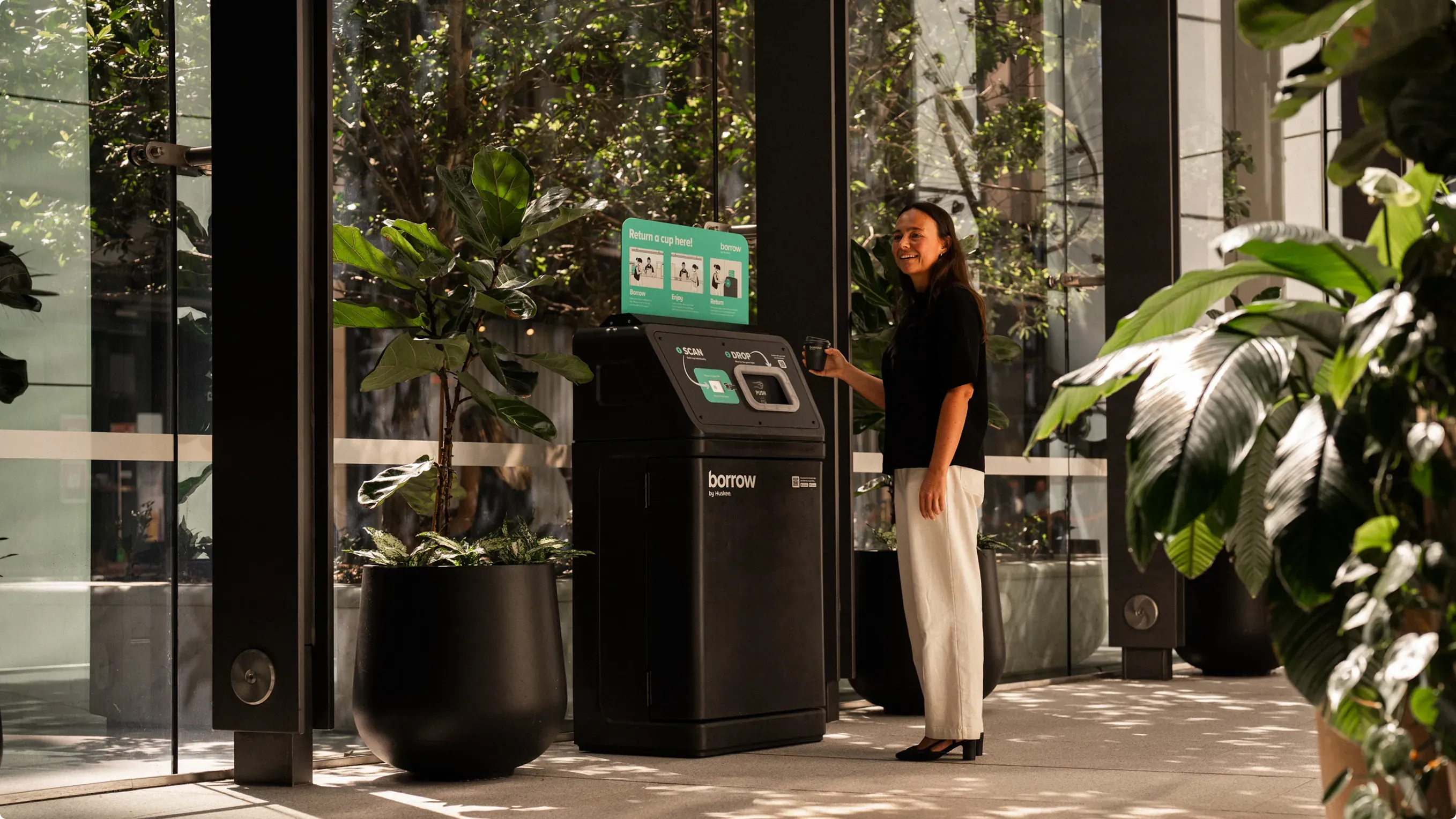 A woman scanning her cup at a Borrow bin in a planted, glass-walled lobby