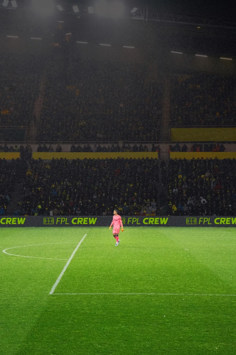 A lone player in pink on a floodlit pitch, CREW hoardings running the length of the touchline
