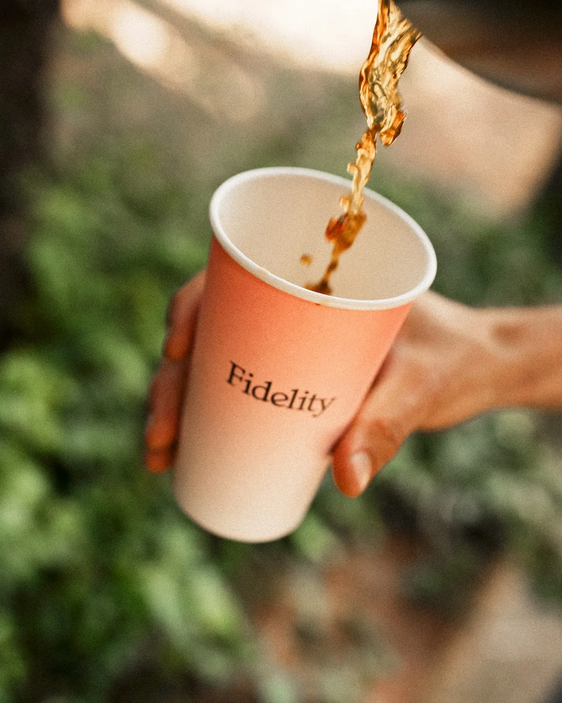 Coffee pouring into a Fidelity branded cup held up against green foliage