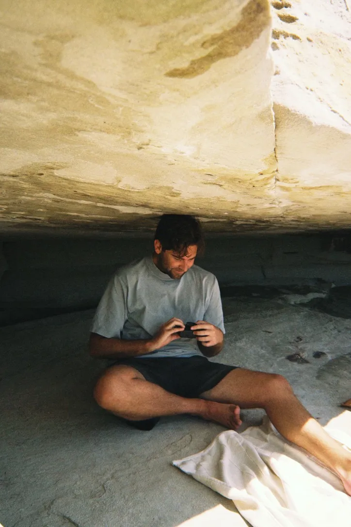 Mark Conway sitting cross-legged under a sandstone overhang, looking at something in his hands