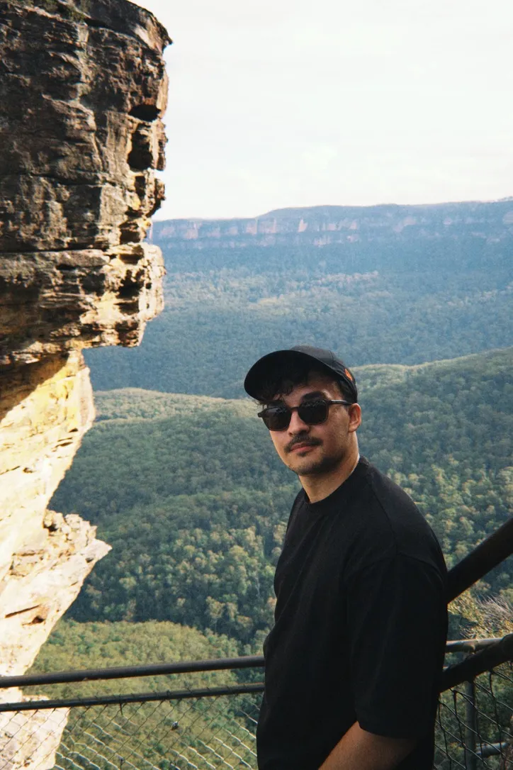 Sam Grimes on a clifftop lookout in a cap and sunglasses, valley and escarpment behind him