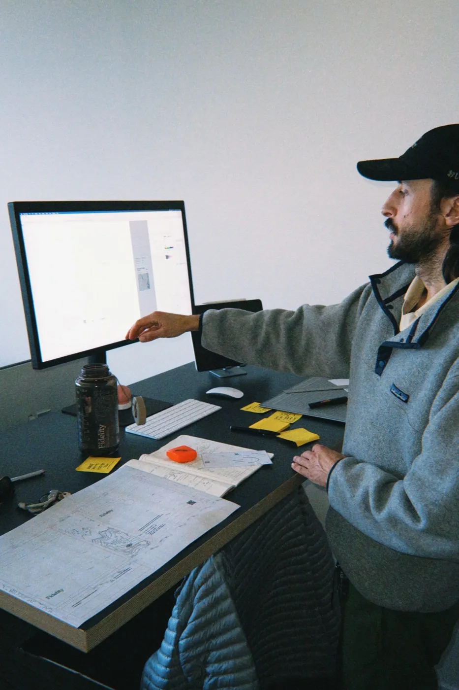 A designer in a cap at a desk, reaching toward a monitor beside printed layouts and sticky notes